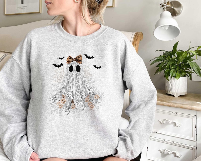 Peut inclure: Un sweat-shirt gris avec un fant&ocirc;me blanc portant un n&oelig;ud brun et entour&eacute; de chauves-souris et de fleurs. Le fant&ocirc;me est fait de dentelle.