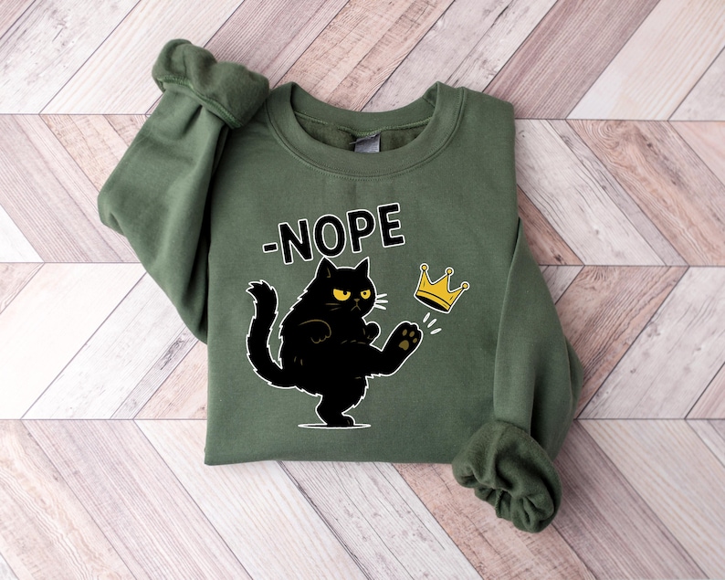 May include: An olive green sweatshirt featuring the word "-NOPE" above a black cat illustration kicking a gold crown. The cat has yellow eyes and white whiskers. The sweatshirt is folded on a light brown and white wooden surface.