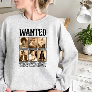 K&ouml;nnte beinhalten: Grauer Pullover mit einem Grafikdruck eines Fahndungsplakats mit Schwarzwei&szlig;fotos von bemerkenswerten Frauen. Der Text auf dem Plakat lautet "WANTED" und "WELL-BEHAVED WOMEN SELDOM MAKE HISTORY".