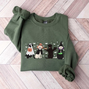May include: An olive green sweatshirt featuring a Halloween-themed graphic. The graphic depicts four dogs dressed in Halloween costumes: a ghost, a witch, a bat, and a witch. The design includes bats and stars.