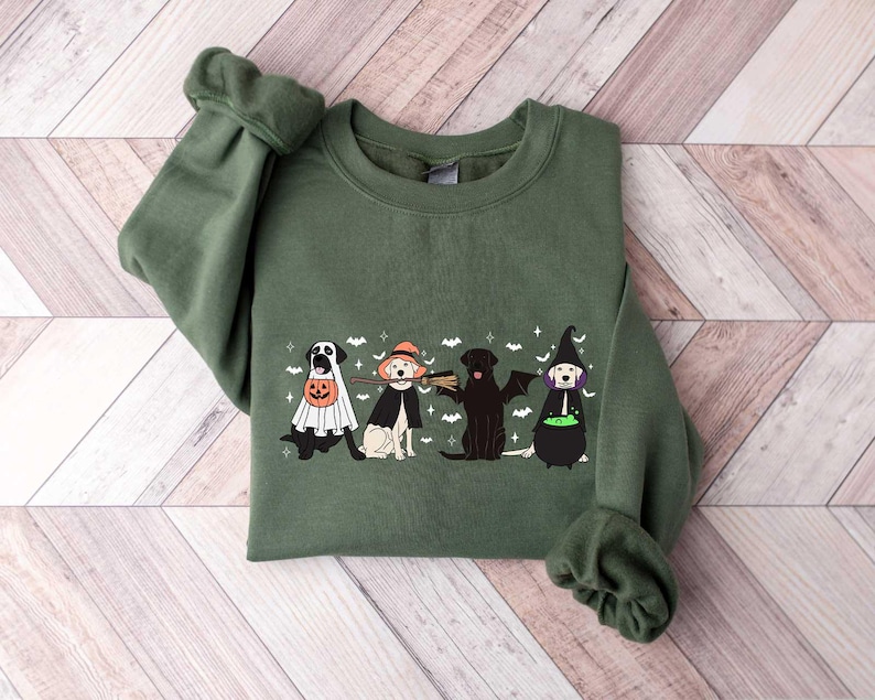 May include: An olive green sweatshirt featuring a Halloween-themed graphic. The graphic depicts four dogs dressed in Halloween costumes: a ghost, a witch, a bat, and a witch. The design includes bats and stars.