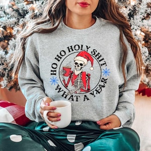 May include: A grey sweatshirt with a black circle graphic featuring a skeleton wearing a Santa hat and holding a mug. The text around the graphic reads "Ho Ho Holy Shit What A Year".