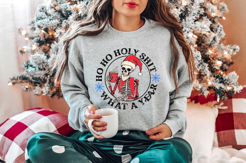 May include: A grey sweatshirt with a black circle graphic featuring a skeleton wearing a Santa hat and holding a mug. The text around the graphic reads "Ho Ho Holy Shit What A Year".