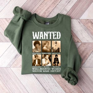 K&ouml;nnte beinhalten: Gr&uuml;ner Pullover mit einem wei&szlig;en Aufdruck, der "WANTED" sagt und Schwarzwei&szlig;fotos von sechs Frauen zeigt. Der Text unter den Fotos lautet "WELL-BEHAVED WOMEN SELDOM MAKE HISTORY."