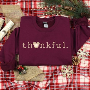 May include: Burgundy sweatshirt with the text "thankful." in a light brown color. A small brown mouse head with a pink bow is in front of the "h" in the word "thankful."