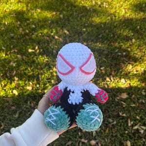 May include: A handmade crochet doll featuring a white head with pink eye accents, a black and white body, and green and black legs. The doll's arms are red and white. The doll is held against a background of green grass and fallen leaves.