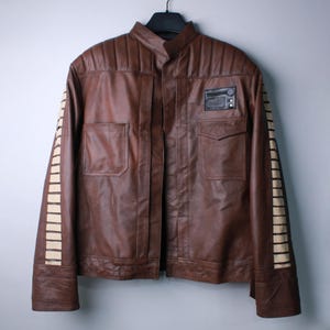 May include: A brown leather jacket featuring a stand-up collar, quilted shoulder panels, and two front pockets. The sleeves are accented with vertical cream-colored rectangular stripes, and a black rectangular patch is attached to the upper chest.