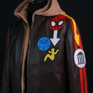 K&ouml;nnte beinhalten: Braune Lederjacke mit beigefarbener Kapuze und mehreren Aufn&auml;hern. Die Jacke hat einen Spider-Man-Aufn&auml;her, einen blauen Sternaufn&auml;her, einen gelben Figurenaufn&auml;her und rote und gelbe Streifen am &Auml;rmel.