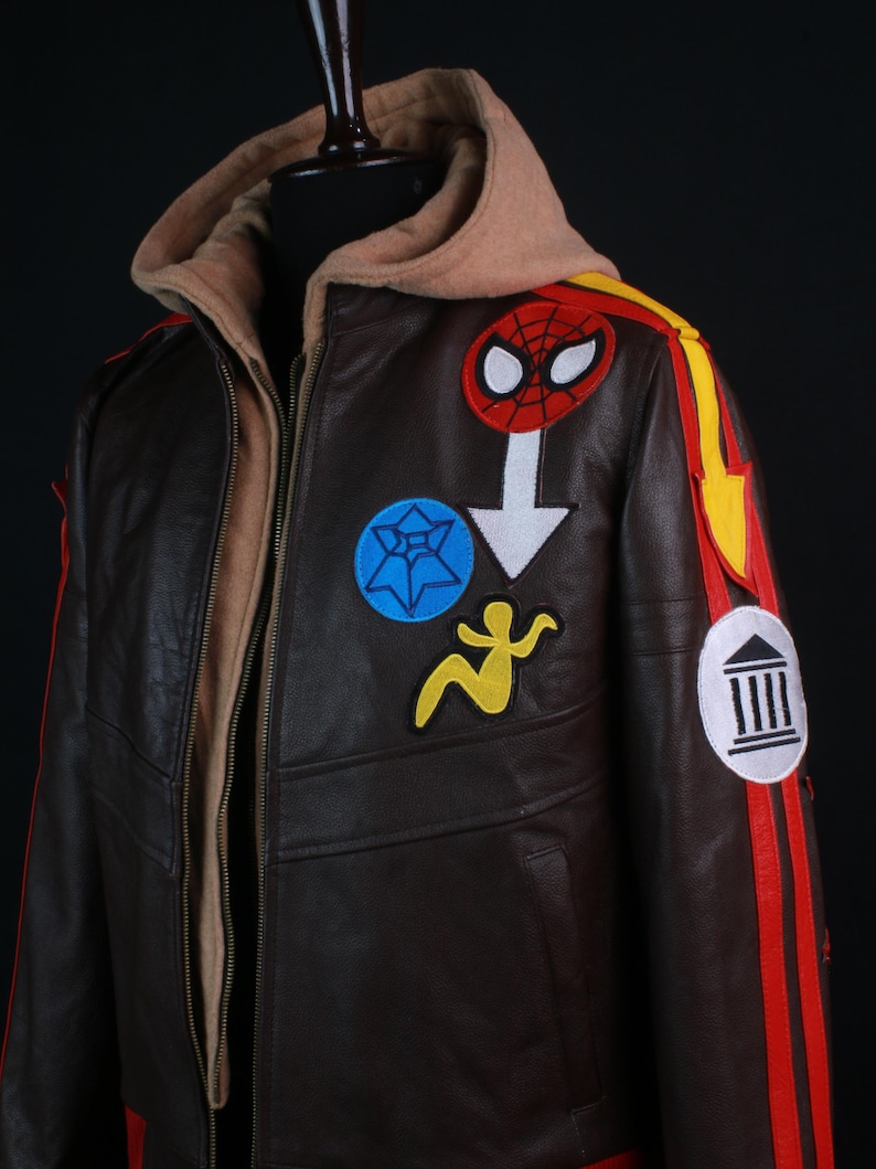 K&ouml;nnte beinhalten: Braune Lederjacke mit beigefarbener Kapuze und mehreren Aufn&auml;hern. Die Jacke hat einen Spider-Man-Aufn&auml;her, einen blauen Sternaufn&auml;her, einen gelben Figurenaufn&auml;her und rote und gelbe Streifen am &Auml;rmel.