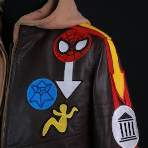 K&ouml;nnte beinhalten: Braune Lederjacke mit beigefarbener Kapuze und mehreren bunten Aufn&auml;hern. Aufn&auml;her sind ein Spider-Man-Logo, ein blauer Stern, eine gelbe Figur und ein wei&szlig;er Pfeil. Rote und gelbe Streifen zieren die Schulter.