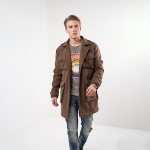 Puede incluir: Un abrigo de lana marrón con múltiples bolsillos y cuello. El abrigo se lleva sobre una camiseta gris con la palabra "GROGU" y un personaje de dibujos animados. La persona lleva vaqueros azules.