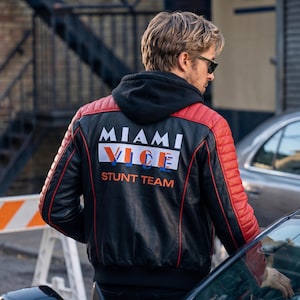 Op de afbeelding: Zwarte leren jas met rode gewatteerde schouders en accenten. De achterkant van de jas toont de tekst "MIAMI VICE STUNT TEAM" in witte, oranje en blauwe letters. De jas heeft een zwarte capuchon.