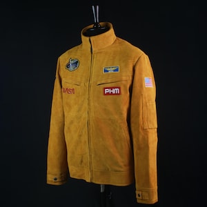 May include: Mustard-colored jacket with a high collar and multiple patches. The jacket features the NASA logo, a small American flag, and the letters "PHM." The jacket has a zipper closure and two front pockets.