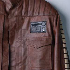 May include: A brown leather jacket featuring a mandarin collar, vertical quilted shoulder panels, and a front chest pocket. A black rectangular patch with a circular detail and grid pattern is positioned above the pocket. The sleeve has a cream stripe.