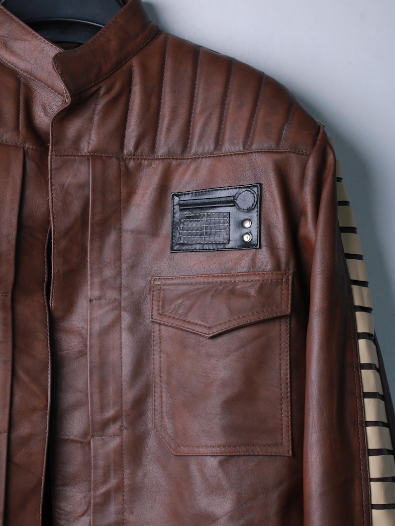 May include: A brown leather jacket featuring a mandarin collar, vertical quilted shoulder panels, and a front chest pocket. A black rectangular patch with a circular detail and grid pattern is positioned above the pocket. The sleeve has a cream stripe.