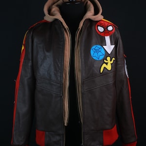 K&ouml;nnte beinhalten: Braune Lederjacke mit beigefarbener Kapuze und roten und gelben Akzenten. Die Jacke hat mehrere Aufn&auml;her, darunter ein Spider-Man-Logo und andere farbenfrohe Designs. Die Manschetten und der untere Saum sind rot.