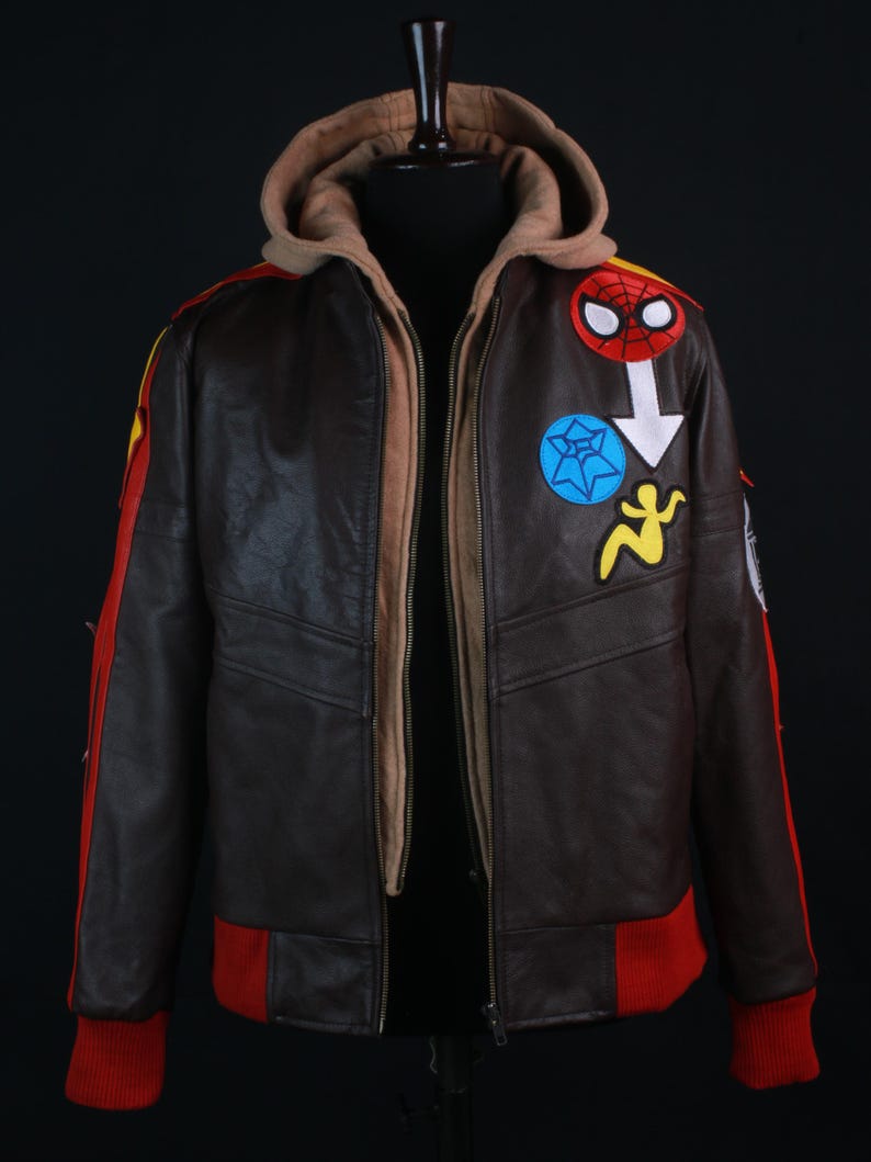 K&ouml;nnte beinhalten: Braune Lederjacke mit beigefarbener Kapuze und roten und gelben Akzenten. Die Jacke hat mehrere Aufn&auml;her, darunter ein Spider-Man-Logo und andere farbenfrohe Designs. Die Manschetten und der untere Saum sind rot.