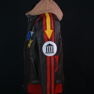 K&ouml;nnte beinhalten: Braune Lederjacke mit beiger Kapuze und roten und gelben Akzenten. Die Jacke hat Aufn&auml;her und einen roten Rippsaum. Ein wei&szlig;er runder Aufn&auml;her mit einem Geb&auml;udedesign befindet sich auf dem &Auml;rmel.