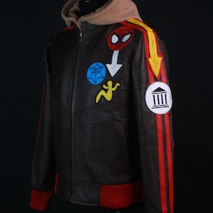 K&ouml;nnte beinhalten: Braune Lederjacke mit beigefarbener Kapuze und roten Akzenten. Die Jacke hat bunte Aufn&auml;her, darunter ein Spider-Man-Emblem und ein gelbes Pfeildesign. Rote Streifen verlaufen die &Auml;rmel hinunter, und die B&uuml;ndchen und der Saum sind rot.