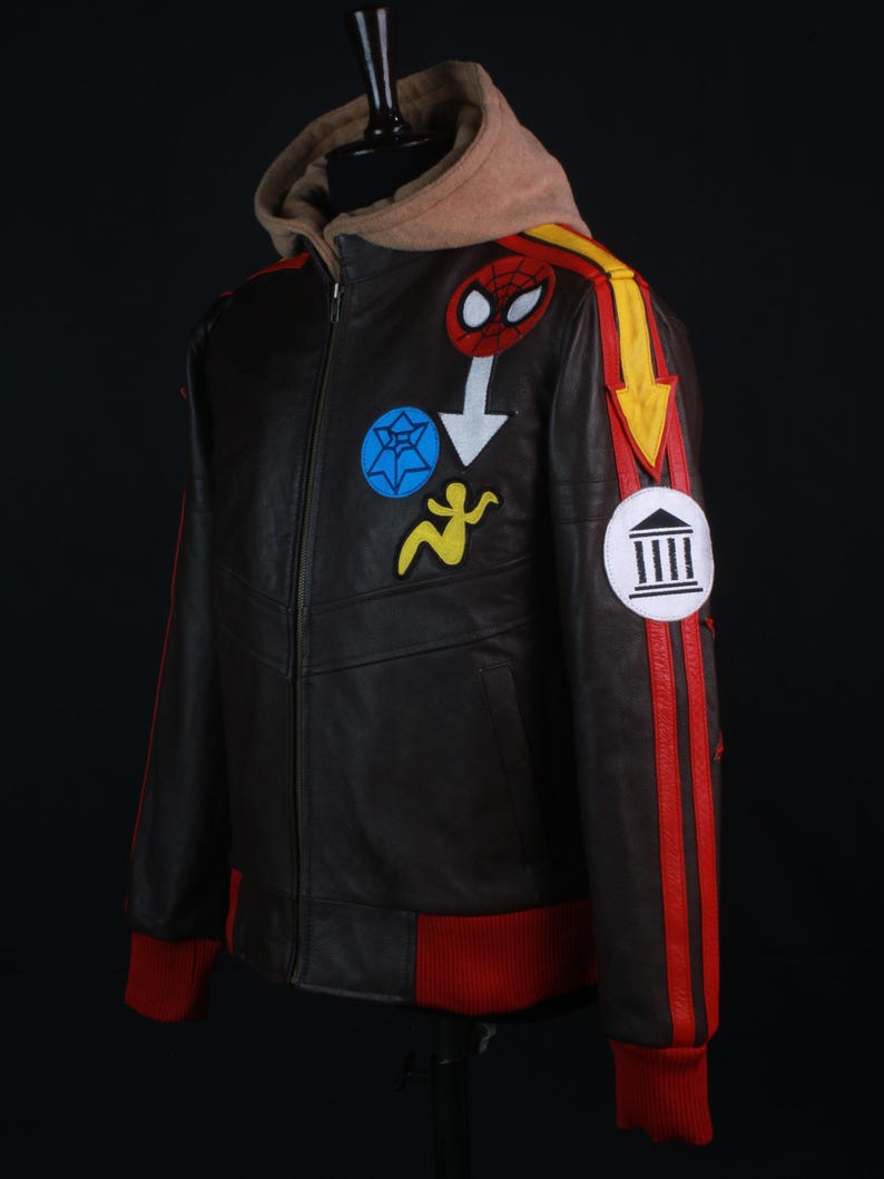 K&ouml;nnte beinhalten: Braune Lederjacke mit beigefarbener Kapuze und roten Akzenten. Die Jacke hat bunte Aufn&auml;her, darunter ein Spider-Man-Emblem und ein gelbes Pfeildesign. Rote Streifen verlaufen die &Auml;rmel hinunter, und die B&uuml;ndchen und der Saum sind rot.