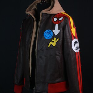 K&ouml;nnte beinhalten: Braune Lederjacke mit beigefarbener Kapuze und roten Akzenten. Die Jacke hat bunte Aufn&auml;her, darunter ein Spider-Man-Logo und andere grafische Designs. Die Manschetten und der Streifen am &Auml;rmel sind rot.