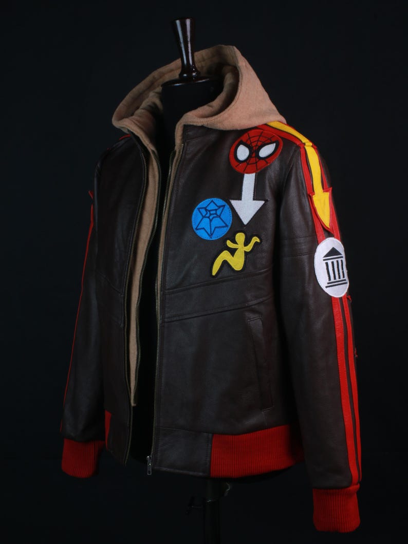 K&ouml;nnte beinhalten: Braune Lederjacke mit beigefarbener Kapuze und roten Akzenten. Die Jacke hat bunte Aufn&auml;her, darunter ein Spider-Man-Logo und andere grafische Designs. Die Manschetten und der Streifen am &Auml;rmel sind rot.