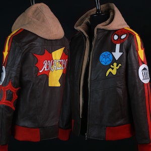 K&ouml;nnte beinhalten: Braune Lederjacke mit beigefarbener Kapuze und roten und gelben Akzenten. Die Jacke hat verschiedene Aufn&auml;her, darunter ein Spinnen-Symbol und das Wort "AMAZIN". Sie hat einen roten Rippensaum und B&uuml;ndchen.