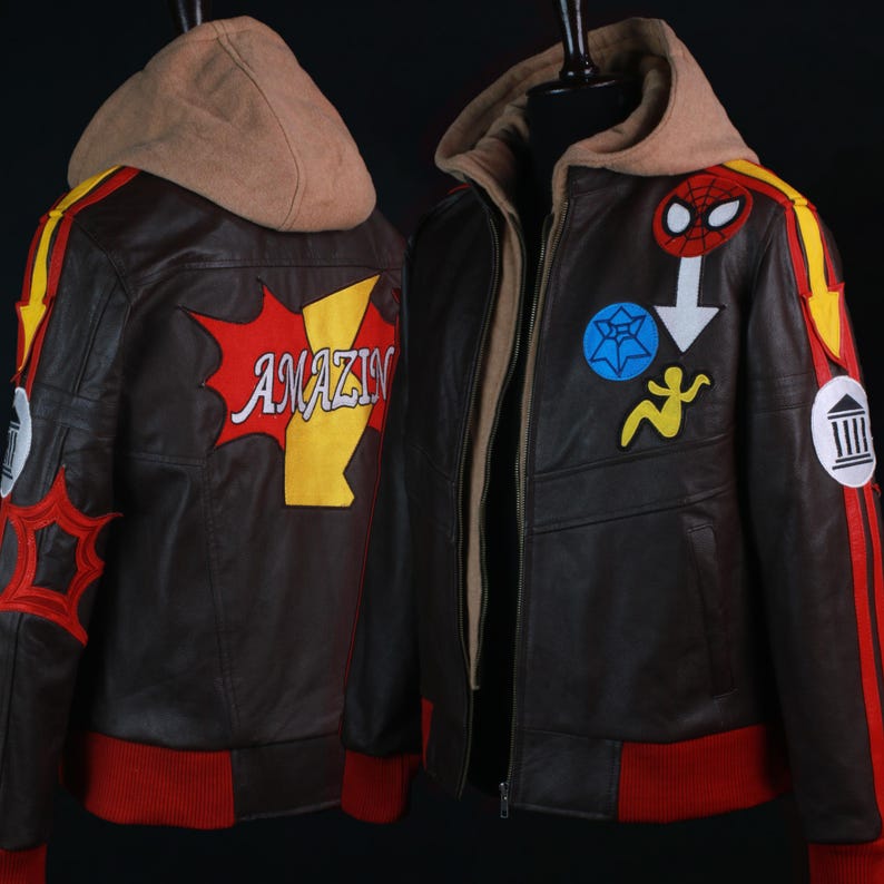 K&ouml;nnte beinhalten: Braune Lederjacke mit beigefarbener Kapuze und roten und gelben Akzenten. Die Jacke hat verschiedene Aufn&auml;her, darunter ein Spinnen-Symbol und das Wort "AMAZIN". Sie hat einen roten Rippensaum und B&uuml;ndchen.