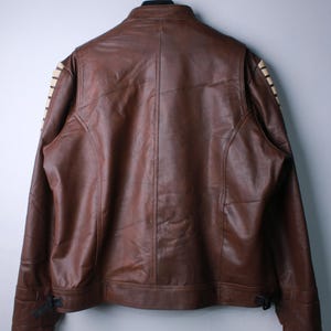 May include: A brown leather jacket featuring a stand-up collar and long sleeves with cream-colored lace-up detailing along the outer arms. The jacket includes a central vertical seam on the back and adjustable buckle straps at the bottom hem.