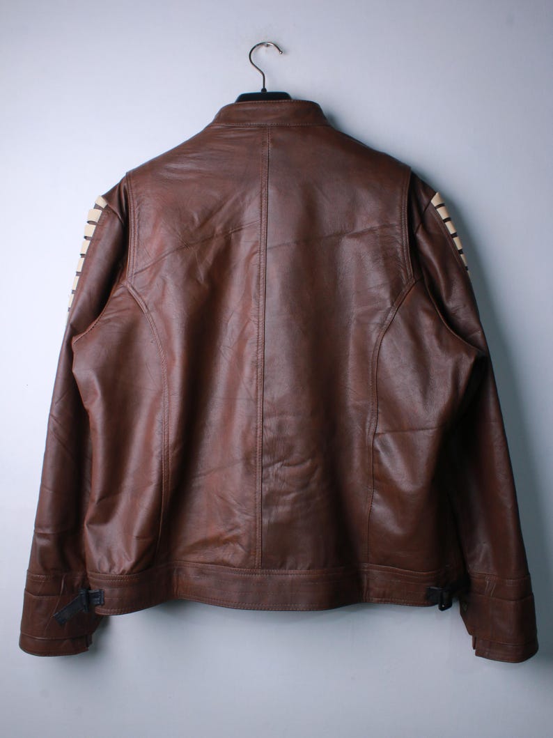 May include: A brown leather jacket featuring a stand-up collar and long sleeves with cream-colored lace-up detailing along the outer arms. The jacket includes a central vertical seam on the back and adjustable buckle straps at the bottom hem.