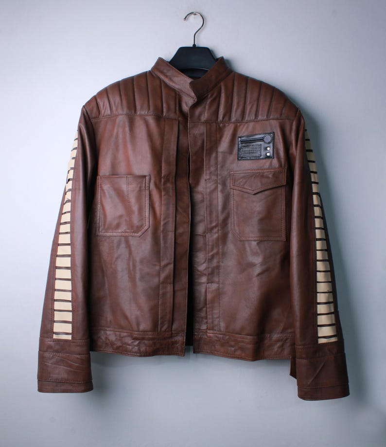 May include: A brown leather jacket featuring a stand-up collar, quilted shoulder panels, and two front pockets. The sleeves are accented with vertical cream-colored rectangular stripes, and a black rectangular patch is attached to the upper chest.