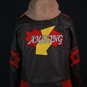 K&ouml;nnte beinhalten: Braune Lederjacke mit beigefarbener Kapuze. Auf dem R&uuml;cken steht das Wort "AMAZING" in Wei&szlig;, ein gelber Blitz und rote Akzente. Rote B&uuml;ndchen und rote spinnennetzartige Designs auf den &Auml;rmeln.