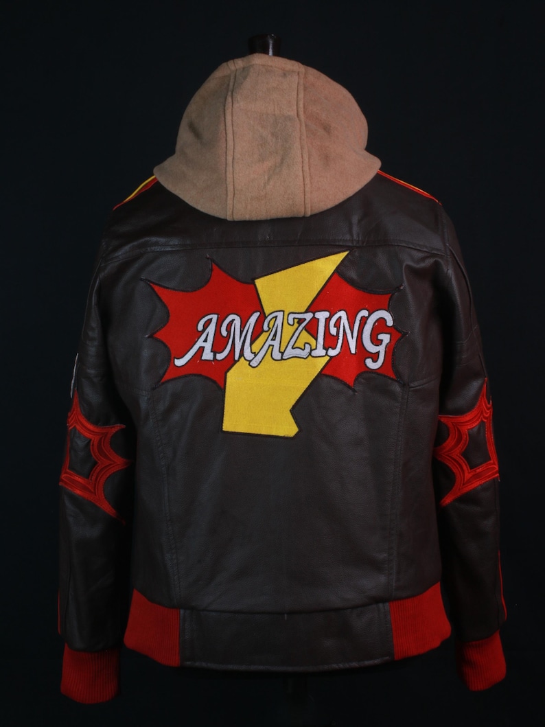 K&ouml;nnte beinhalten: Braune Lederjacke mit beigefarbener Kapuze. Auf dem R&uuml;cken steht das Wort "AMAZING" in Wei&szlig;, ein gelber Blitz und rote Akzente. Rote B&uuml;ndchen und rote spinnennetzartige Designs auf den &Auml;rmeln.