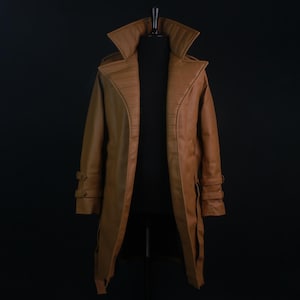 May include: A tan trench coat featuring a wide, structured collar with horizontal stitching, long sleeves with adjustable buckled straps at the cuffs, and a smooth finish. The coat is displayed on a mannequin against a solid black background.