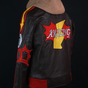 K&ouml;nnte beinhalten: Braune Lederjacke mit beigefarbener Kapuze und roten und gelben Akzenten. Auf der R&uuml;ckseite der Jacke steht das Wort "AMAZING" in einem Comic-Design. Die Jacke hat rote B&uuml;ndchen und einen roten Saum.