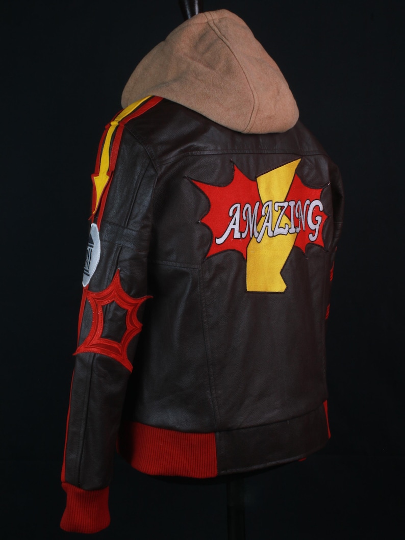 K&ouml;nnte beinhalten: Braune Lederjacke mit beigefarbener Kapuze und roten und gelben Akzenten. Auf der R&uuml;ckseite der Jacke steht das Wort "AMAZING" in einem Comic-Design. Die Jacke hat rote B&uuml;ndchen und einen roten Saum.