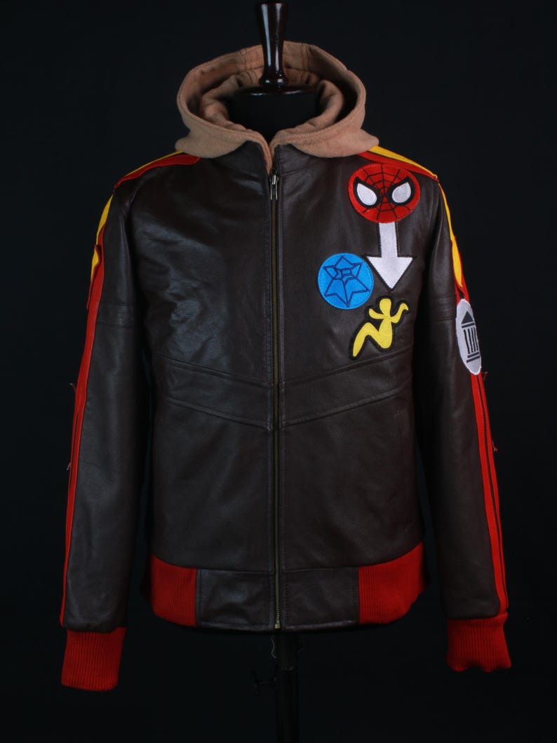 K&ouml;nnte beinhalten: Braune Lederjacke mit beigefarbener Kapuze und roten und gelben Akzenten. Die Jacke hat einen Spider-Man-Aufn&auml;her und andere bunte gestickte Designs. Die B&uuml;ndchen und der Bund sind rot, und die Jacke hat einen durchgehenden Rei&szlig;verschluss.