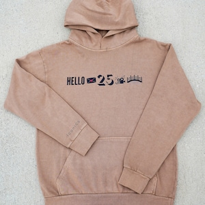 May include: A brown hooded sweatshirt with the text "HELLO 25" and a few small icons printed on the front. The text "iconics" is printed on the left sleeve.