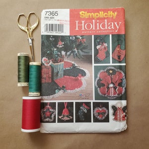 May include: A Simplicity Holiday pattern collection book featuring a Christmas tree skirt, stocking, and ornaments. The book is open to reveal the patterns and instructions. There are three spools of thread in green and red, and a pair of gold scissors.