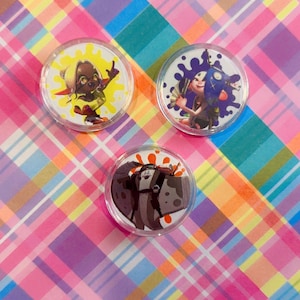 May include: Three circular acrylic charms featuring colorful character illustrations from the Splatoon video game series. Each charm displays a unique character against a vibrant paint splatter background, encased in a clear, protective circular frame.