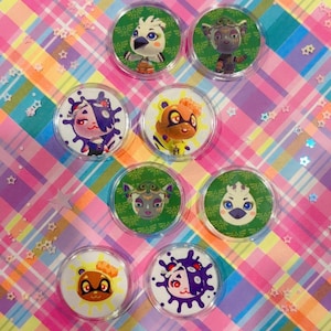 May include: A collection of eight circular, clear-backed pins featuring colorful cartoon character designs. The pins are arranged on a vibrant, multicolored plaid background with star-shaped confetti.