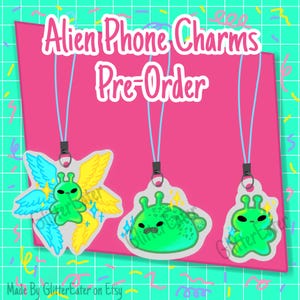 May include: Three green alien phone charms with blue lanyards against a pink backdrop. The charms have varied designs, including one with wings. The text "Alien Phone Charms Pre-Order" is displayed at the top of the image.