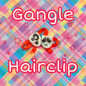 Puede incluir: Una pinza para el pelo con dos pequeñas máscaras blancas con rasgos faciales negros, una sonriente y otra triste, unidas a un lazo de cinta roja brillante. El texto Gangle Hairclip aparece en letras blancas grandes sobre un fondo de cuadros.