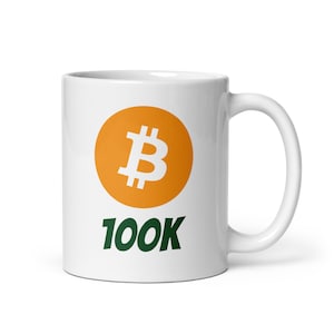 Puede incluir: Taza de cerámica blanca con un círculo naranja grande que contiene un símbolo de Bitcoin blanco. El texto "100K" está escrito en verde debajo del círculo.