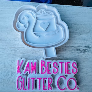 May include: White silicone mold of a flamingo holding a can with a heart on it. Below the mold is a pink glitter sign that reads "KAM BESTIES GLITTER CO."