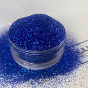 Op de afbeelding: Een kleine, doorzichtige plastic pot gevuld met helderblauwe glitter. De glitter is verspreid rond de pot op een wit oppervlak. De glitter heeft een sprankelend effect en reflecteert licht. De pot is gelabeld met de tekst "A.M. BLANKS".