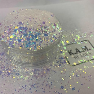 Può includere: Un contenitore di plastica trasparente pieno di glitter iridescenti. I glitter sono un mix di bianco, blu e viola, con una qualità scintillante e riflettente. Il contenitore è etichettato "K.A.M. BLANKS".