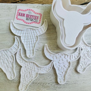 Puede incluir: Cinco moldes de cráneo de toro de resina blanca. Los moldes tienen diferentes patrones, incluyendo una cuadrícula, formas geométricas y un diseño floral. Un molde es un molde de dos piezas para un cráneo de toro más grande.