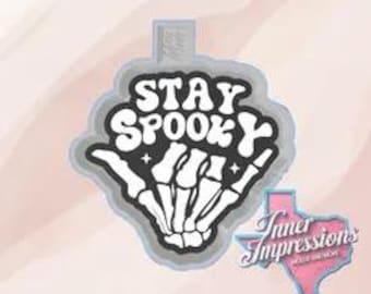 May include: A gray and white cookie cutter in the shape of a hand with the text "STAY SPOOKY". The hand is a skeleton hand. A pink Texas-shaped logo with the text "Inner Impressions" is in the lower right corner.