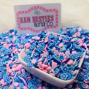 Puede incluir: Un tazón blanco lleno de espolvoreados coloridos. Los espolvoreados tienen forma de flores y palitos pequeños. Los espolvoreados son de color rosa, azul, morado y blanco. El tazón está sobre una superficie blanca. El texto "KAM BESTIES GLITTER CO." es visible en una etiqueta rosa y blanca.